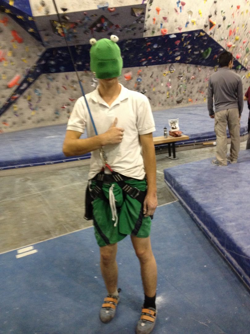 A Rock Climber in a Climbing Gym Wearing a Frog Hat Over His Face
